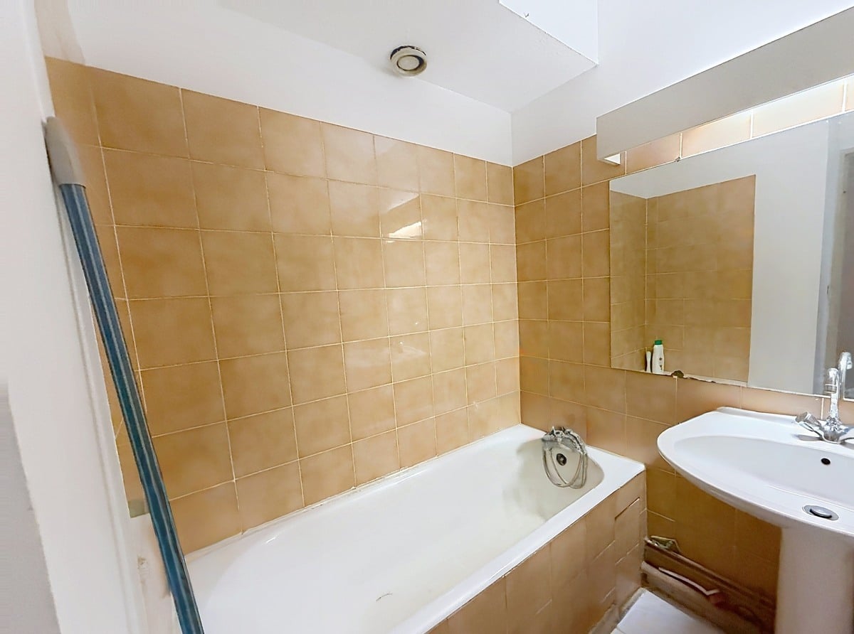 Salle de bain entièrement carrelée de grands carreaux beige-ocre brillants, équipée d'une baignoire encastrée blanche avec mitigeur chromé et d'un lavabo vasque blanc sur meuble bois arrondi. Un miroir rectangulaire sans cadre est fixé au-dessus du lavabo, et une ventilation mécanique circulaire est visible au plafond blanc.