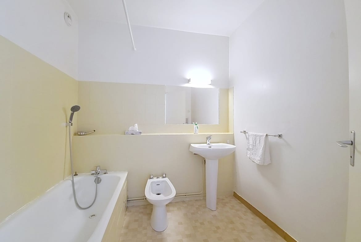 Salle de bains rectangulaire avec une baignoire encastrée équipée d'un flexible de douche chromé, un bidet blanc au sol et un lavabo sur colonne. Les murs sont peints en blanc au-dessus d'un soubassement jaune clair, surmontés d'un large miroir rectangulaire éclairé par une applique murale.