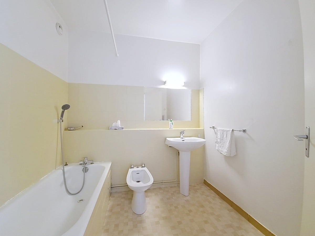 Salle de bains rectangulaire avec une baignoire encastrée équipée d'un flexible de douche chromé, un bidet blanc au sol et un lavabo sur colonne. Les murs sont peints en blanc au-dessus d'un soubassement jaune clair, surmontés d'un large miroir rectangulaire éclairé par une applique murale.