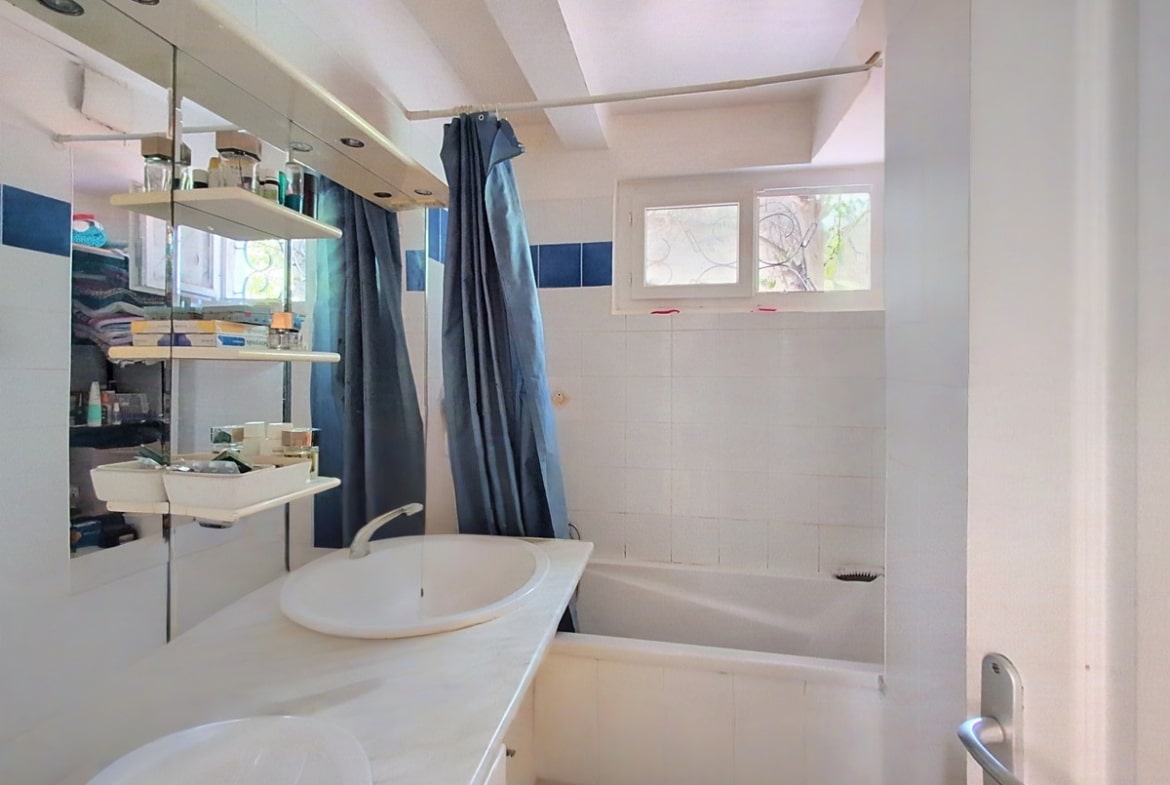 Salle de bain carrelée blanc et bleu avec vasque ronde blanche posée sur plan, miroir à éclairage intégré et étagères murales chromées chargées de produits. Baignoire encastrée avec rideau de douche bleu marine et deux fenêtres à imposte apportant la lumière naturelle.