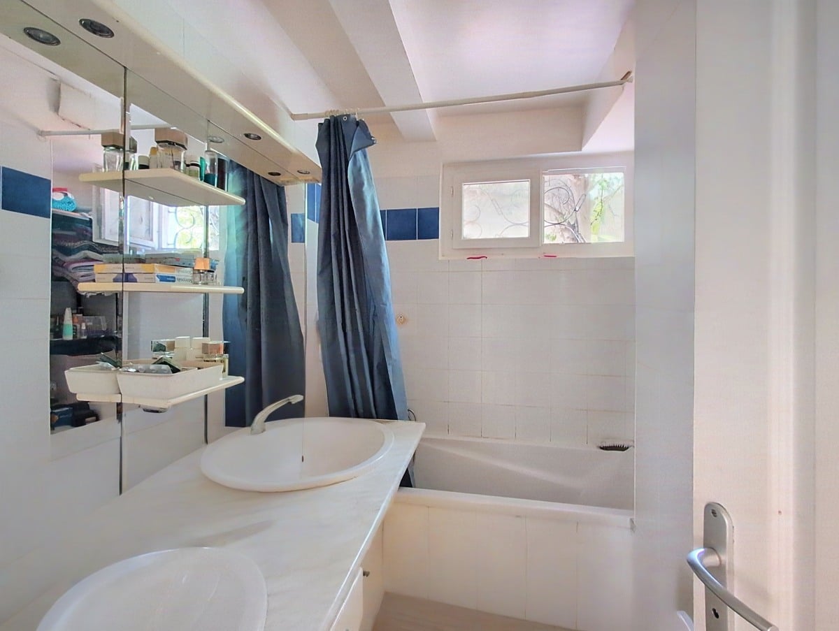 Salle de bain carrelée blanc et bleu avec vasque ronde blanche posée sur plan, miroir à éclairage intégré et étagères murales chromées chargées de produits. Baignoire encastrée avec rideau de douche bleu marine et deux fenêtres à imposte apportant la lumière naturelle.