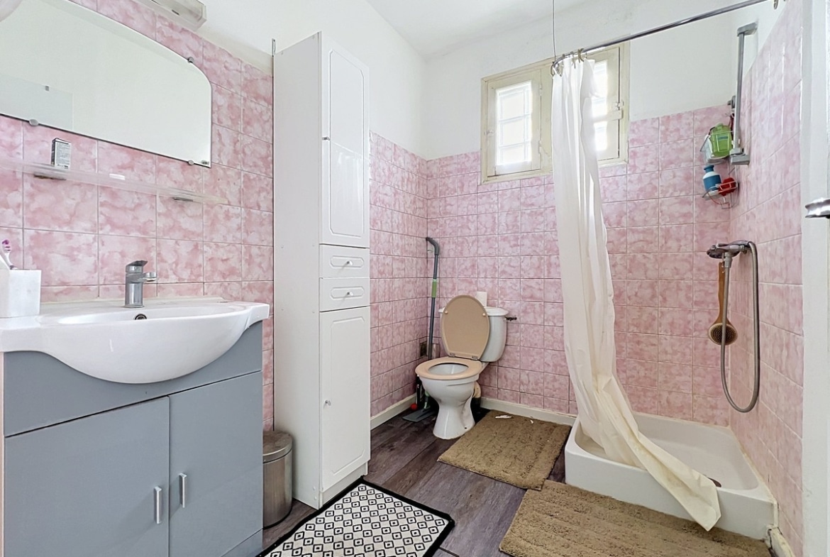 Salle de bain avec WC intégré entièrement carrelée de faïences roses à effet marbré, équipée d'un lavabo vasque blanc sur meuble gris, d'une colonne de rangement blanche et d'un bac à douche avec rideau blanc. Petite fenêtre à battants donnant sur l'extérieur, sol en revêtement imitation parquet gris, tapis géométrique noir et blanc au sol.