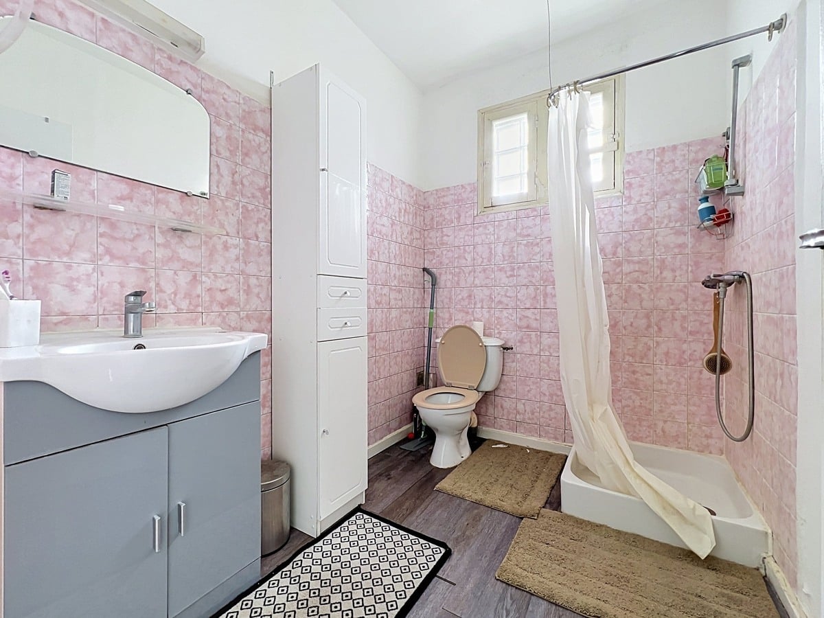 Salle de bain avec WC intégré entièrement carrelée de faïences roses à effet marbré, équipée d'un lavabo vasque blanc sur meuble gris, d'une colonne de rangement blanche et d'un bac à douche avec rideau blanc. Petite fenêtre à battants donnant sur l'extérieur, sol en revêtement imitation parquet gris, tapis géométrique noir et blanc au sol.