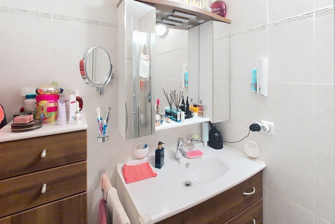 Salle de bain carrelée de blanc avec un meuble vasque suspendu en bois foncé surmonté d'un miroir à trois panneaux avec éclairage intégré et rangements latéraux. Un meuble à tiroirs en bois assorti est positionné à gauche du lavabo, avec divers produits de soin et accessoires disposés sur les surfaces.