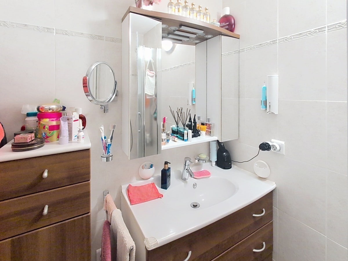 Salle de bain carrelée de blanc avec un meuble vasque suspendu en bois foncé surmonté d'un miroir à trois panneaux avec éclairage intégré et rangements latéraux. Un meuble à tiroirs en bois assorti est positionné à gauche du lavabo, avec divers produits de soin et accessoires disposés sur les surfaces.