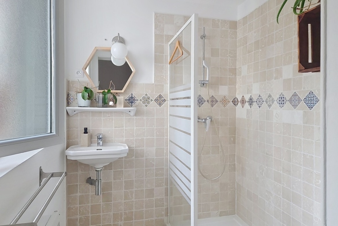 Salle de bain avec revêtement mural en carreaux beige clair et frise de carreaux motifs bleus en losange, cabine de douche à paroi vitrée avec colonne de douche chromée. Lave-mains suspendu blanc, miroir hexagonal encadré de bois clair et sèche-serviettes blanc fixé au mur.