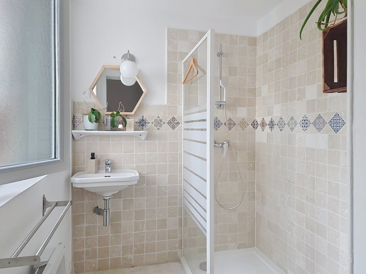 Salle de bain avec revêtement mural en carreaux beige clair et frise de carreaux motifs bleus en losange, cabine de douche à paroi vitrée avec colonne de douche chromée. Lave-mains suspendu blanc, miroir hexagonal encadré de bois clair et sèche-serviettes blanc fixé au mur.