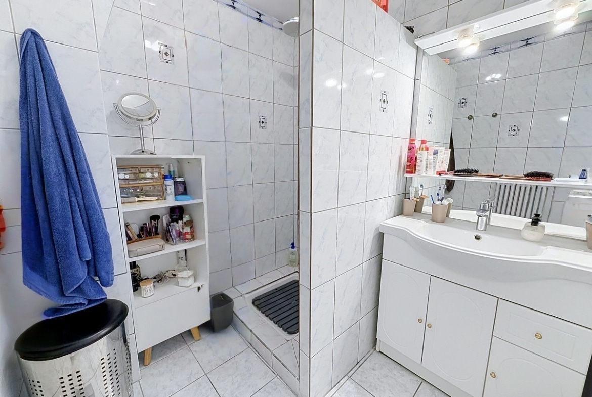 Salle de bain carrelée de faïence blanche effet marbre avec douche à l'italienne équipée d'un receveur bas et meuble vasque blanc à deux portes surmonté d'un miroir éclairé. Petite étagère de rangement blanche, serviette bleue accrochée et corbeille à linge au sol.