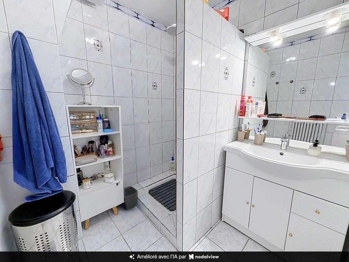 Salle de bain carrelée de faïence blanche effet marbre avec douche à l'italienne équipée d'un receveur bas et meuble vasque blanc à deux portes surmonté d'un miroir éclairé. Petite étagère de rangement blanche, serviette bleue accrochée et corbeille à linge au sol.
