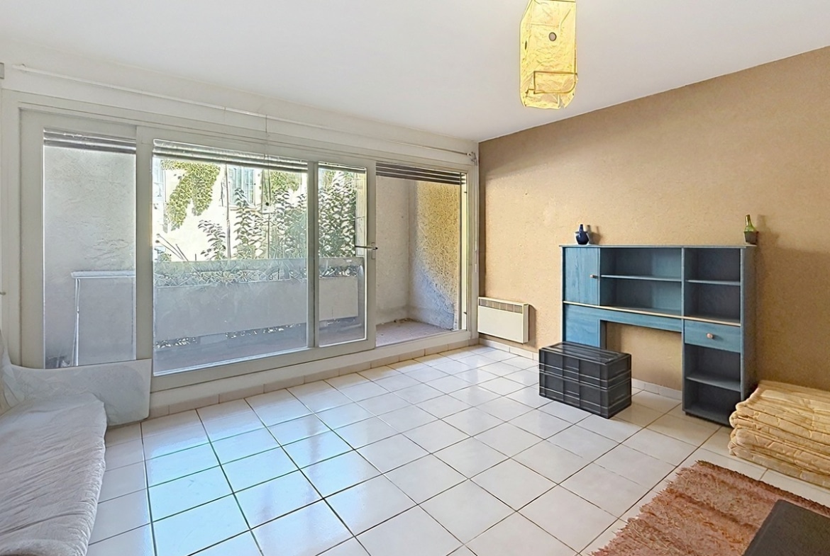 Séjour carrelé de grands carreaux blancs avec baie vitrée coulissante donnant directement sur la terrasse végétalisée, un radiateur électrique blanc fixé sous la baie et un mur d'accent en enduit beige-brun. Un meuble étagère bleu vintage avec tiroir et porte est positionné contre le mur droit, accompagné d'une malle en plastique noir et d'un tapis rouge au sol.