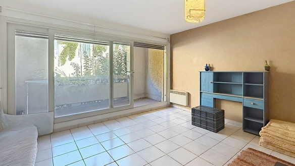 Séjour carrelé de grands carreaux blancs avec baie vitrée coulissante donnant directement sur la terrasse végétalisée, un radiateur électrique blanc fixé sous la baie et un mur d'accent en enduit beige-brun. Un meuble étagère bleu vintage avec tiroir et porte est positionné contre le mur droit, accompagné d'une malle en plastique noir et d'un tapis rouge au sol.