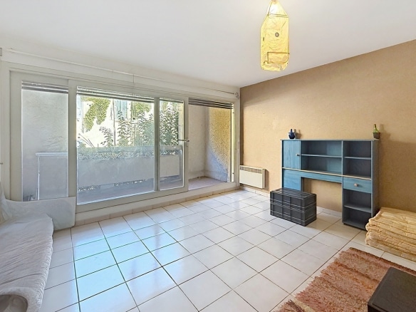 Séjour carrelé de grands carreaux blancs avec baie vitrée coulissante donnant directement sur la terrasse végétalisée, un radiateur électrique blanc fixé sous la baie et un mur d'accent en enduit beige-brun. Un meuble étagère bleu vintage avec tiroir et porte est positionné contre le mur droit, accompagné d'une malle en plastique noir et d'un tapis rouge au sol.