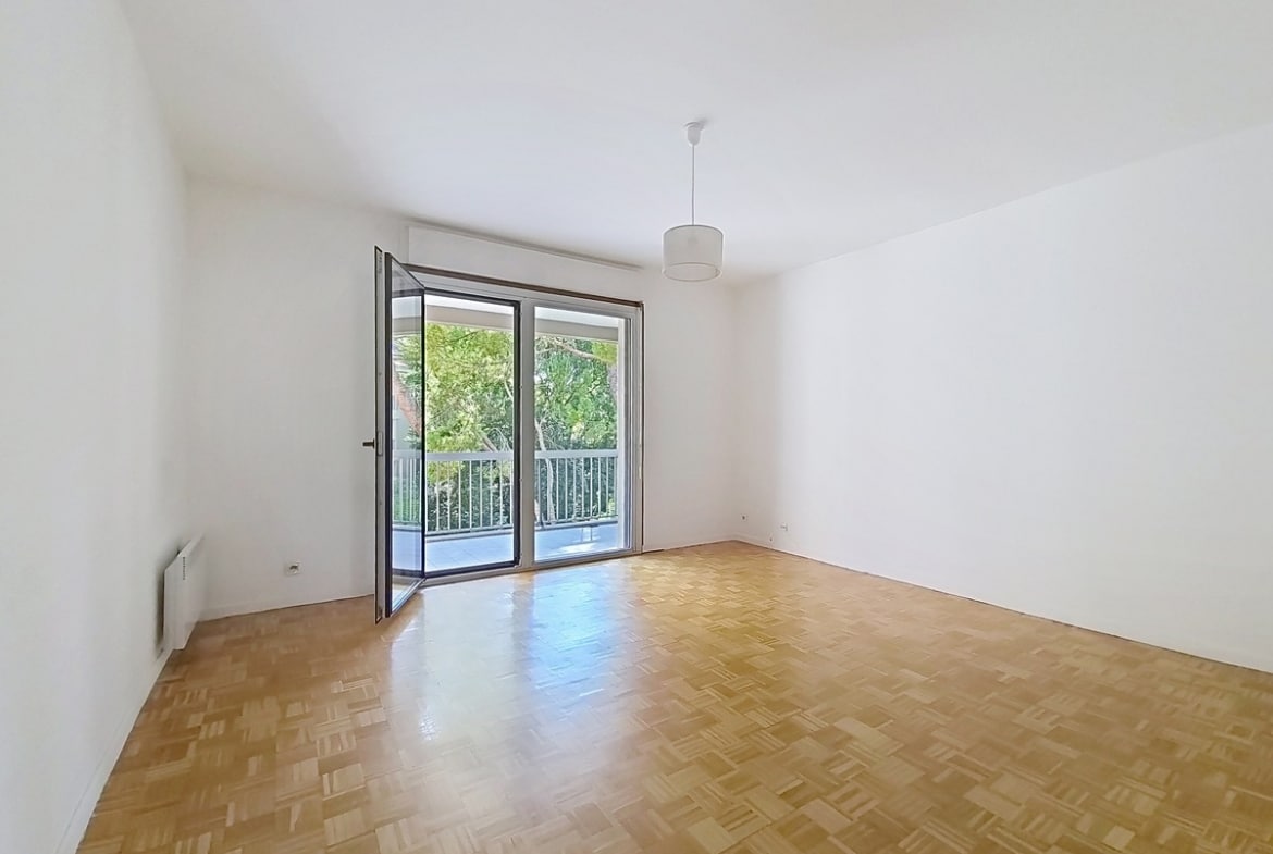 Pièce de séjour vide avec un parquet en point de Hongrie aux tons dorés et des murs blancs, équipée d'un convecteur électrique et d'un plafonnier central. Une large porte-fenêtre à double vantail ouverte donne directement accès au balcon avec vue sur des arbres.