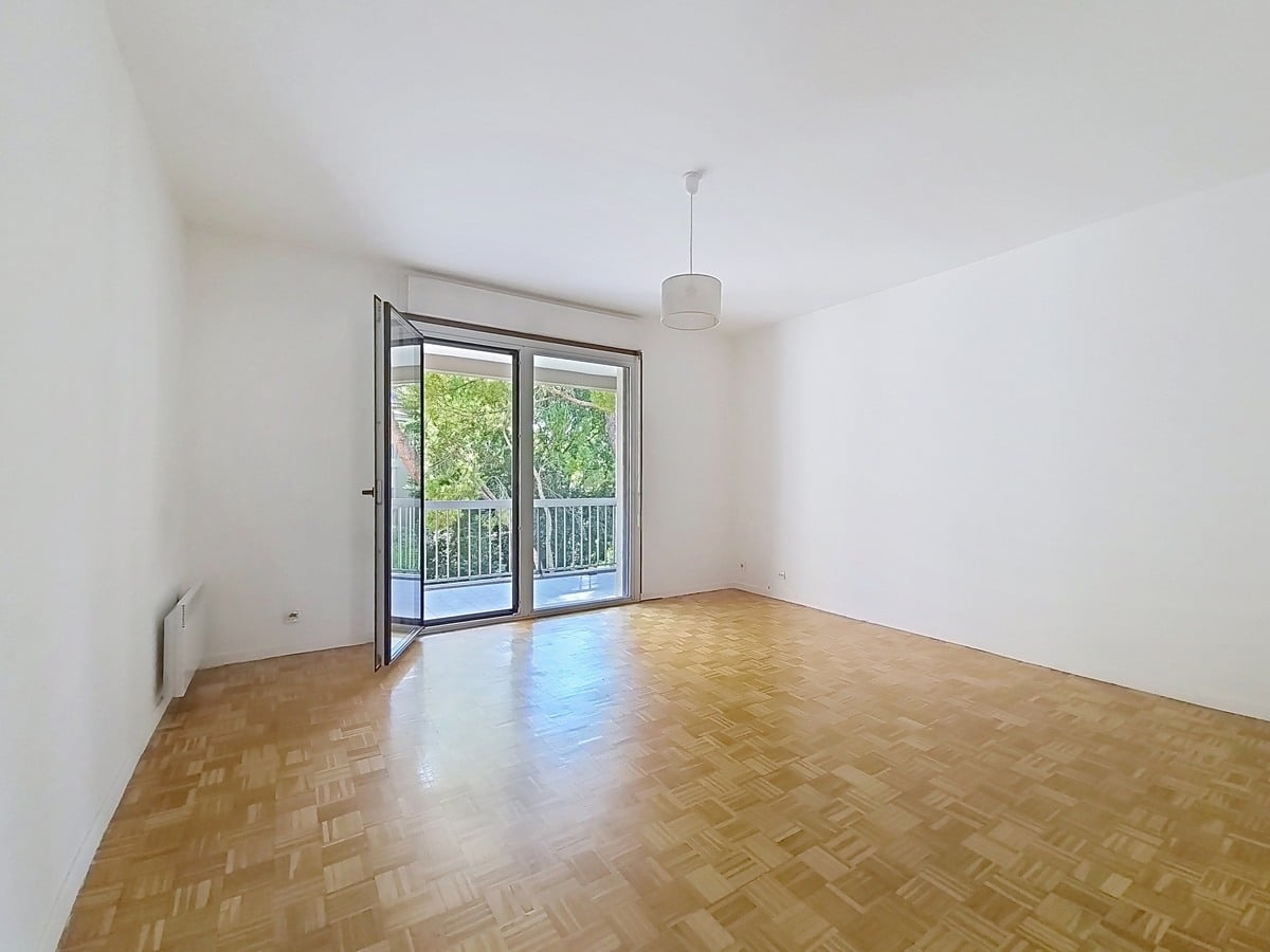 Pièce de séjour vide avec un parquet en point de Hongrie aux tons dorés et des murs blancs, équipée d'un convecteur électrique et d'un plafonnier central. Une large porte-fenêtre à double vantail ouverte donne directement accès au balcon avec vue sur des arbres.