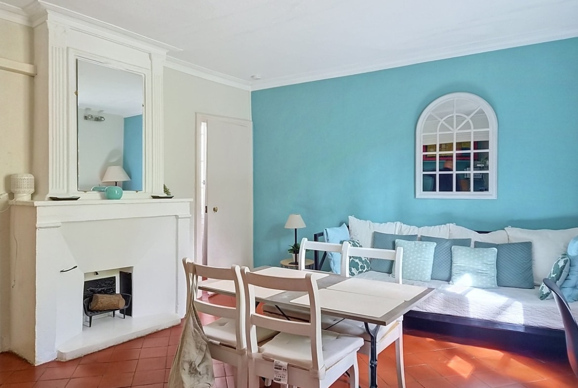 Pièce de vie avec mur peint en bleu turquoise, cheminée blanche surmontée d'un miroir rectangulaire et d'objets décoratifs, sol en tomettes rouges. Table à manger blanche avec quatre chaises en bois clair, canapé recouvert de coussins bleus et blancs longeant le mur du fond orné d'un miroir en forme d'arche.