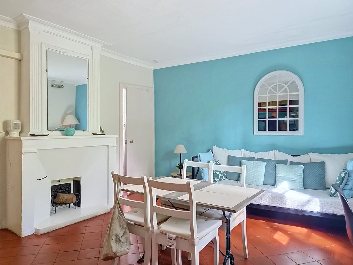 Pièce de vie avec mur peint en bleu turquoise, cheminée blanche surmontée d'un miroir rectangulaire et d'objets décoratifs, sol en tomettes rouges. Table à manger blanche avec quatre chaises en bois clair, canapé recouvert de coussins bleus et blancs longeant le mur du fond orné d'un miroir en forme d'arche.