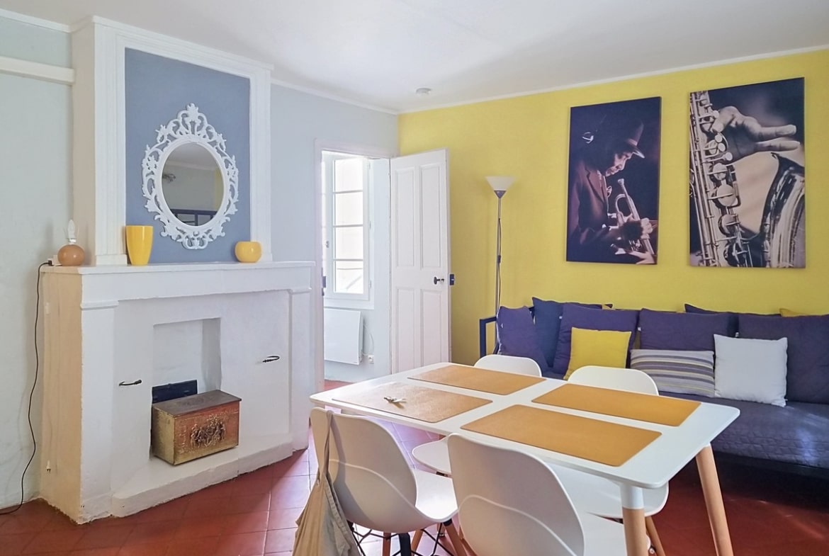 Pièce de vie avec mur d'accent jaune vif orné de deux tableaux photographiques en noir et blanc représentant des musiciens de jazz, cheminée blanche avec encadrement à pilastres et miroir baroque blanc au-dessus. Table à manger blanche avec sets de table ocre et quatre chaises blanches à pieds bois, canapé bleu marine garni de coussins jaunes et gris, sol en tomettes rouges.