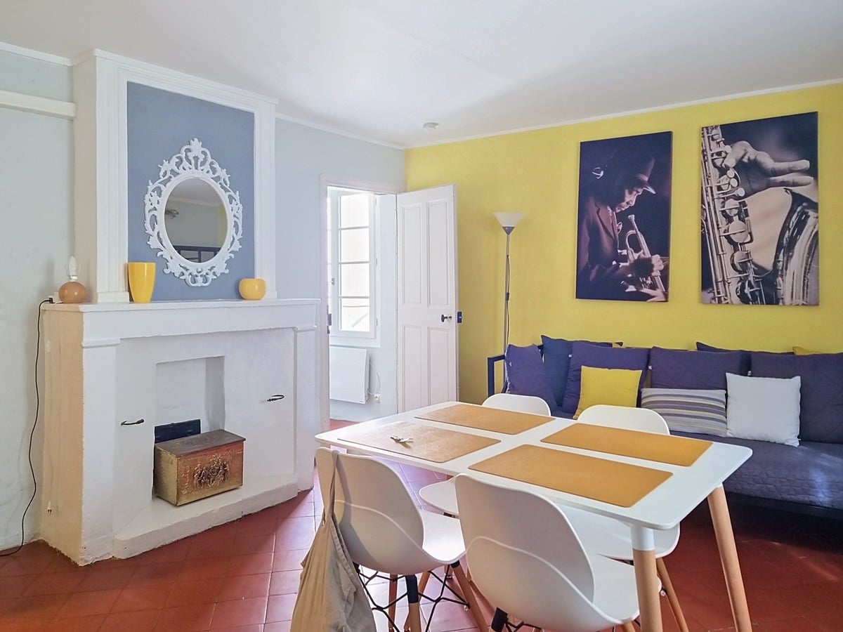 Pièce de vie avec mur d'accent jaune vif orné de deux tableaux photographiques en noir et blanc représentant des musiciens de jazz, cheminée blanche avec encadrement à pilastres et miroir baroque blanc au-dessus. Table à manger blanche avec sets de table ocre et quatre chaises blanches à pieds bois, canapé bleu marine garni de coussins jaunes et gris, sol en tomettes rouges.