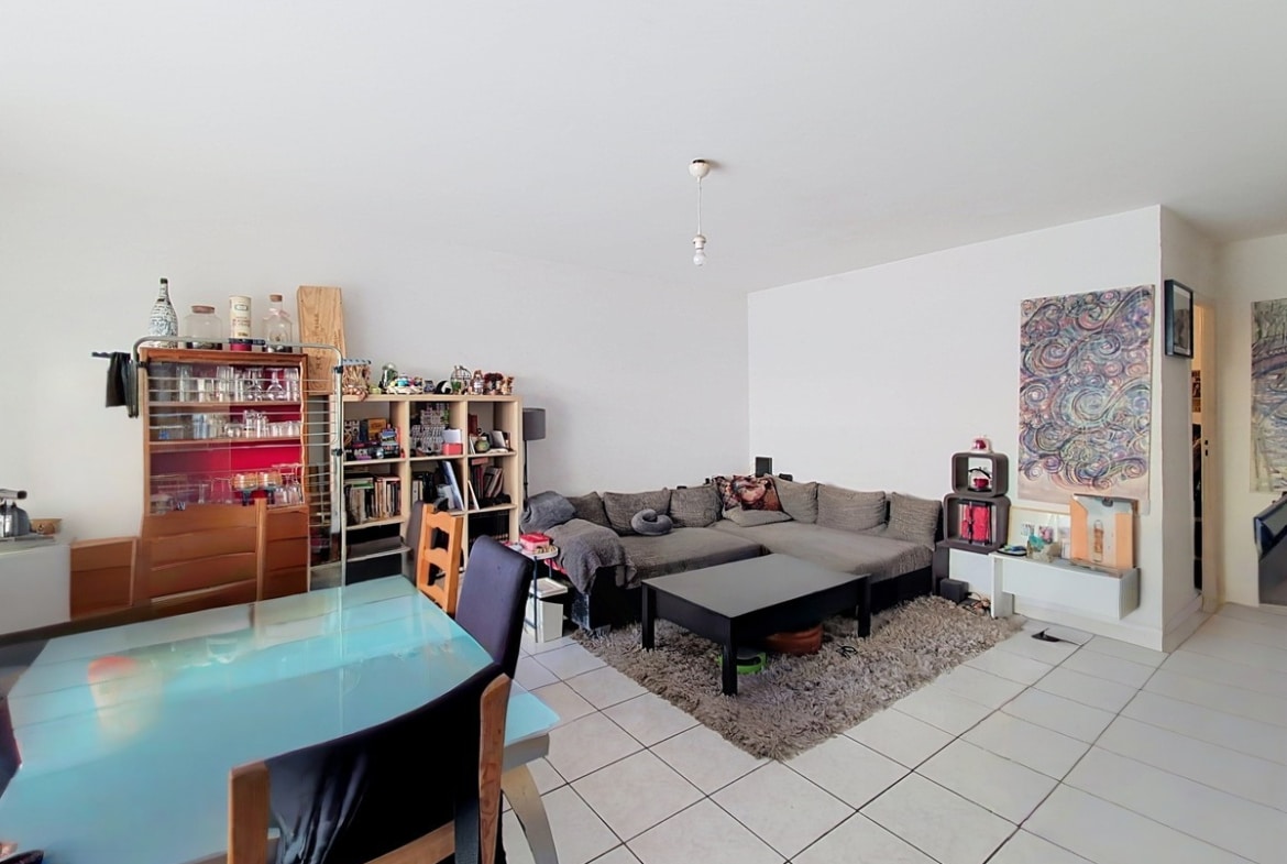 Pièce de vie avec table à manger en verre bleu turquoise et chaises en bois, face à un canapé d'angle gris posé sur un tapis beige à poils longs. Bibliothèques et meubles de rangement en bois occupent le fond de la pièce, avec un tableau coloré accroché au mur blanc.