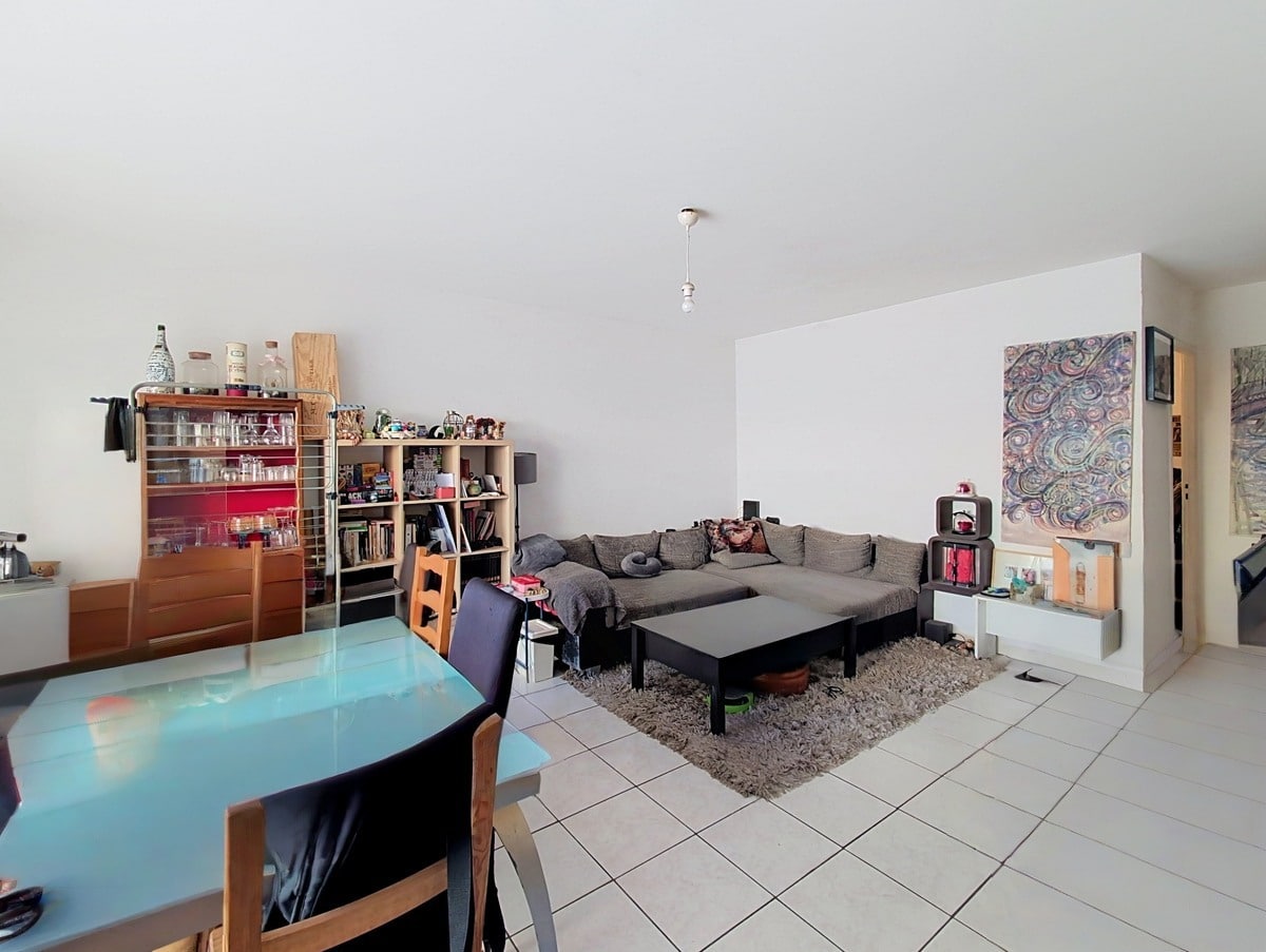 Pièce de vie avec table à manger en verre bleu turquoise et chaises en bois, face à un canapé d'angle gris posé sur un tapis beige à poils longs. Bibliothèques et meubles de rangement en bois occupent le fond de la pièce, avec un tableau coloré accroché au mur blanc.