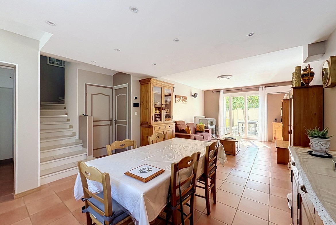 Pièce de vie ouverte avec table à manger rectangulaire recouverte d'une nappe blanche entourée de chaises en bois, donnant sur un salon avec canapé marron. Escalier blanc visible en fond, carrelage tomette rouge au sol et baie vitrée donnant sur la terrasse.