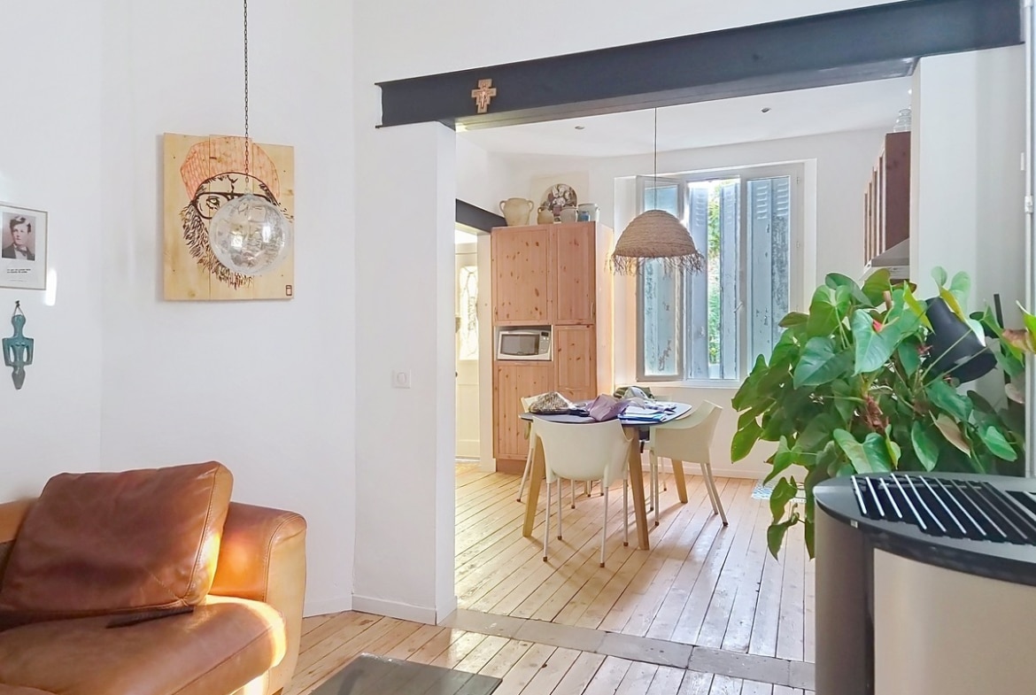 Pièce de vie avec canapé en cuir marron vintage, parquet bois clair et double hauteur sous plafond blanc, ouverte sur la salle à manger via un passage structuré d'une poutre apparente peinte en noir. Tableau sur bois représentant un visage sur le mur blanc, plante verte de grande taille au premier plan.