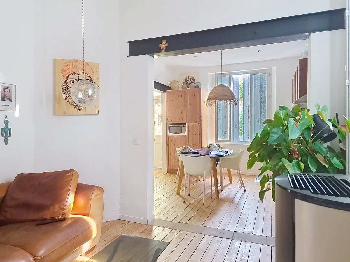 Pièce de vie avec canapé en cuir marron vintage, parquet bois clair et double hauteur sous plafond blanc, ouverte sur la salle à manger via un passage structuré d'une poutre apparente peinte en noir. Tableau sur bois représentant un visage sur le mur blanc, plante verte de grande taille au premier plan.