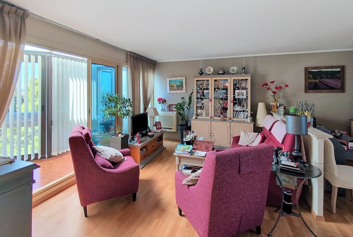 Salon-séjour avec parquet clair et murs taupe, meublé de deux grands fauteuils en tissu rose foncé, d'un buffet vitrine beige et d'un ensemble table basse et téléviseur. Une large baie vitrée à lames verticales ouvre sur une terrasse vitrée baignée de lumière naturelle avec vue sur la verdure.