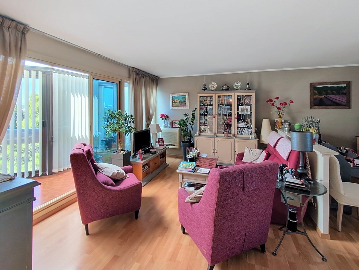 Salon-séjour avec parquet clair et murs taupe, meublé de deux grands fauteuils en tissu rose foncé, d'un buffet vitrine beige et d'un ensemble table basse et téléviseur. Une large baie vitrée à lames verticales ouvre sur une terrasse vitrée baignée de lumière naturelle avec vue sur la verdure.