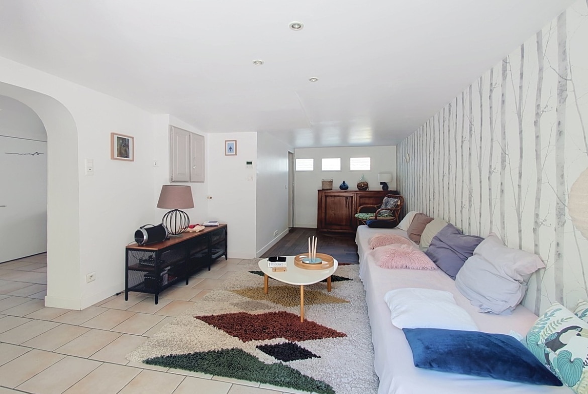 Grande pièce de vie en longueur avec sol en carrelage beige, équipée d'un canapé d'angle blanc recouvert de coussins roses et gris, posé sur un tapis shaggy multicolore. Le mur droit est tapissé d'un papier peint motif bouleau blanc, et une arche en plâtre délimite l'entrée côté gauche.