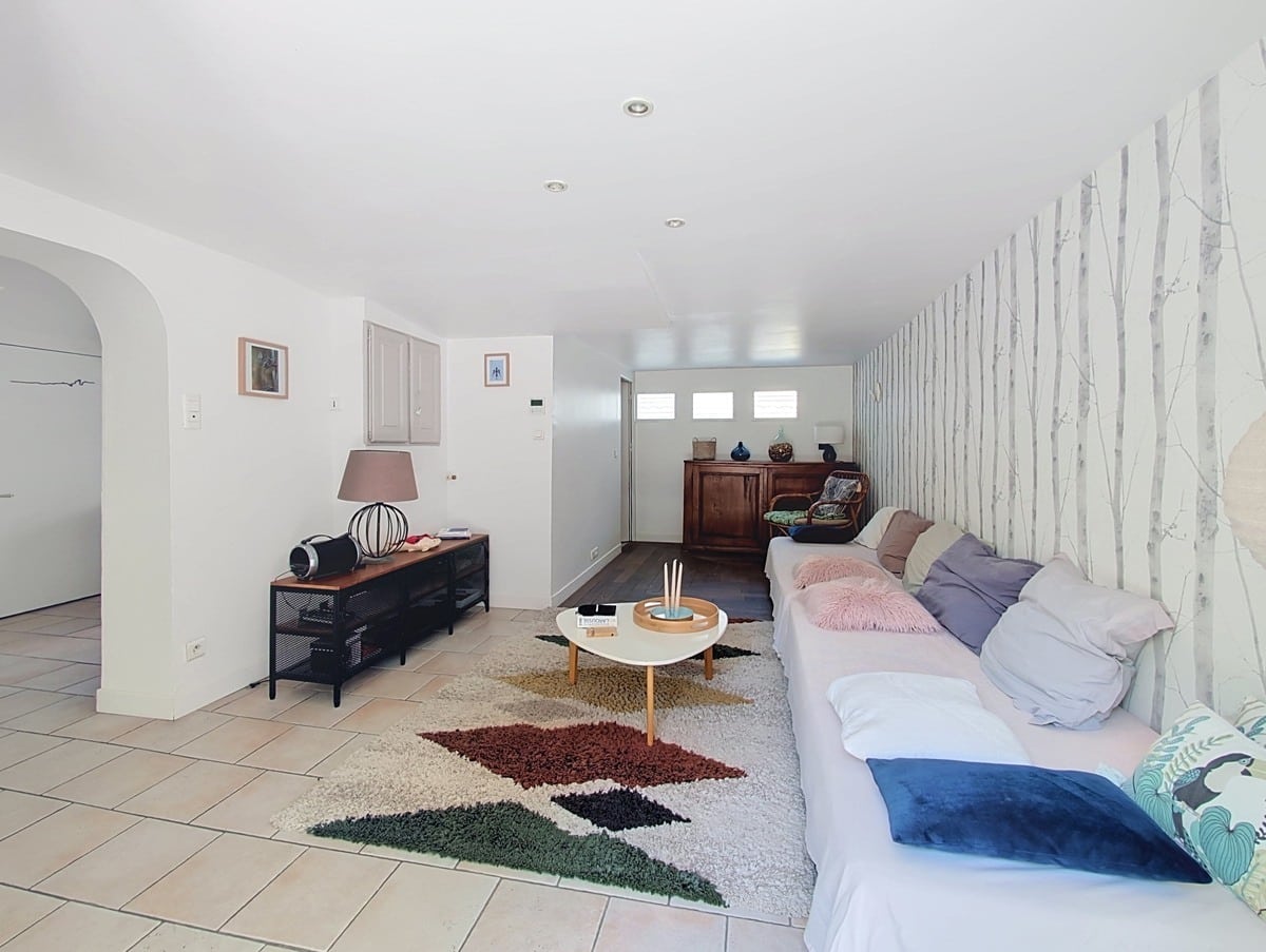 Grande pièce de vie en longueur avec sol en carrelage beige, équipée d'un canapé d'angle blanc recouvert de coussins roses et gris, posé sur un tapis shaggy multicolore. Le mur droit est tapissé d'un papier peint motif bouleau blanc, et une arche en plâtre délimite l'entrée côté gauche.
