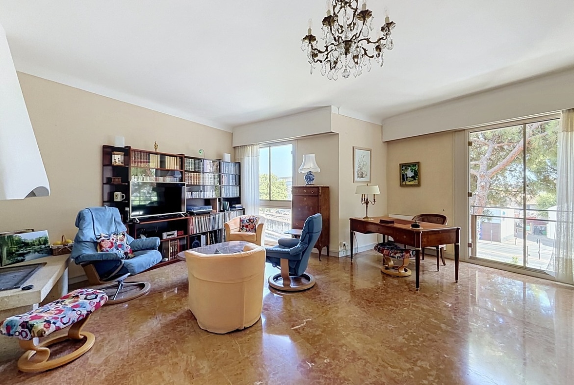 Salon rectangulaire avec sol en marbre poli, éclairé par deux baies vitrées coulissantes donnant sur des balcons et des arbres, meublé d'un ensemble de fauteuils beige et bleu, d'un bureau en bois et d'une vitrine en bois massif contenant de la vaisselle. Un lustre en bronze et cristal est suspendu au plafond blanc.
