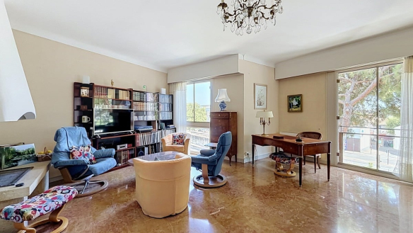 Salon rectangulaire avec sol en marbre poli, éclairé par deux baies vitrées coulissantes donnant sur des balcons et des arbres, meublé d'un ensemble de fauteuils beige et bleu, d'un bureau en bois et d'une vitrine en bois massif contenant de la vaisselle. Un lustre en bronze et cristal est suspendu au plafond blanc.