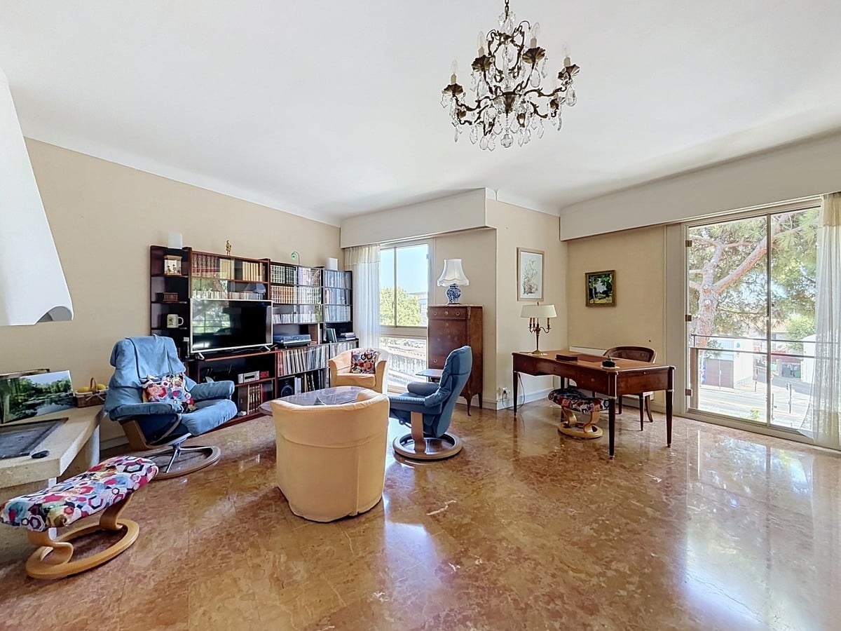 Salon rectangulaire avec sol en marbre poli, éclairé par deux baies vitrées coulissantes donnant sur des balcons et des arbres, meublé d'un ensemble de fauteuils beige et bleu, d'un bureau en bois et d'une vitrine en bois massif contenant de la vaisselle. Un lustre en bronze et cristal est suspendu au plafond blanc.