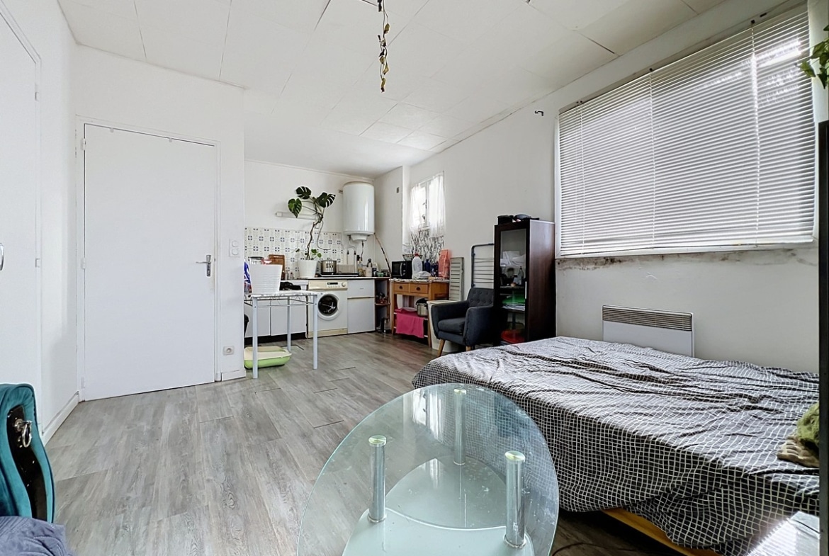 Vue d'ensemble du studio depuis le coin séjour vers la cuisine, montrant la continuité de l'espace unique avec sol en revêtement imitation parquet clair et porte d'entrée blanche à gauche. Lit double avec dessus-de-lit gris en premier plan à droite, table basse ovale en verre fumé au centre, murs blancs et fenêtre à jalousie horizontale apportant la lumière naturelle.