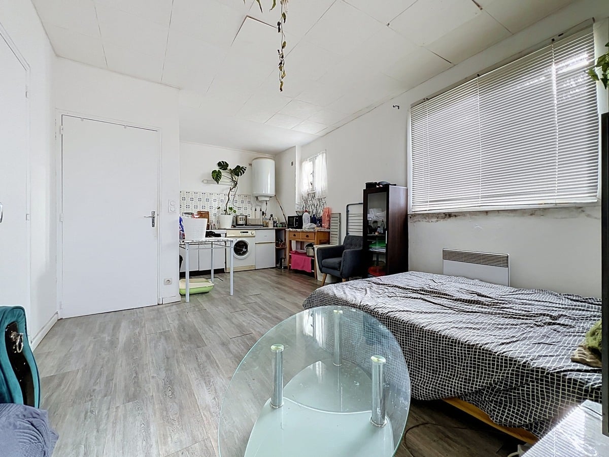Vue d'ensemble du studio depuis le coin séjour vers la cuisine, montrant la continuité de l'espace unique avec sol en revêtement imitation parquet clair et porte d'entrée blanche à gauche. Lit double avec dessus-de-lit gris en premier plan à droite, table basse ovale en verre fumé au centre, murs blancs et fenêtre à jalousie horizontale apportant la lumière naturelle.