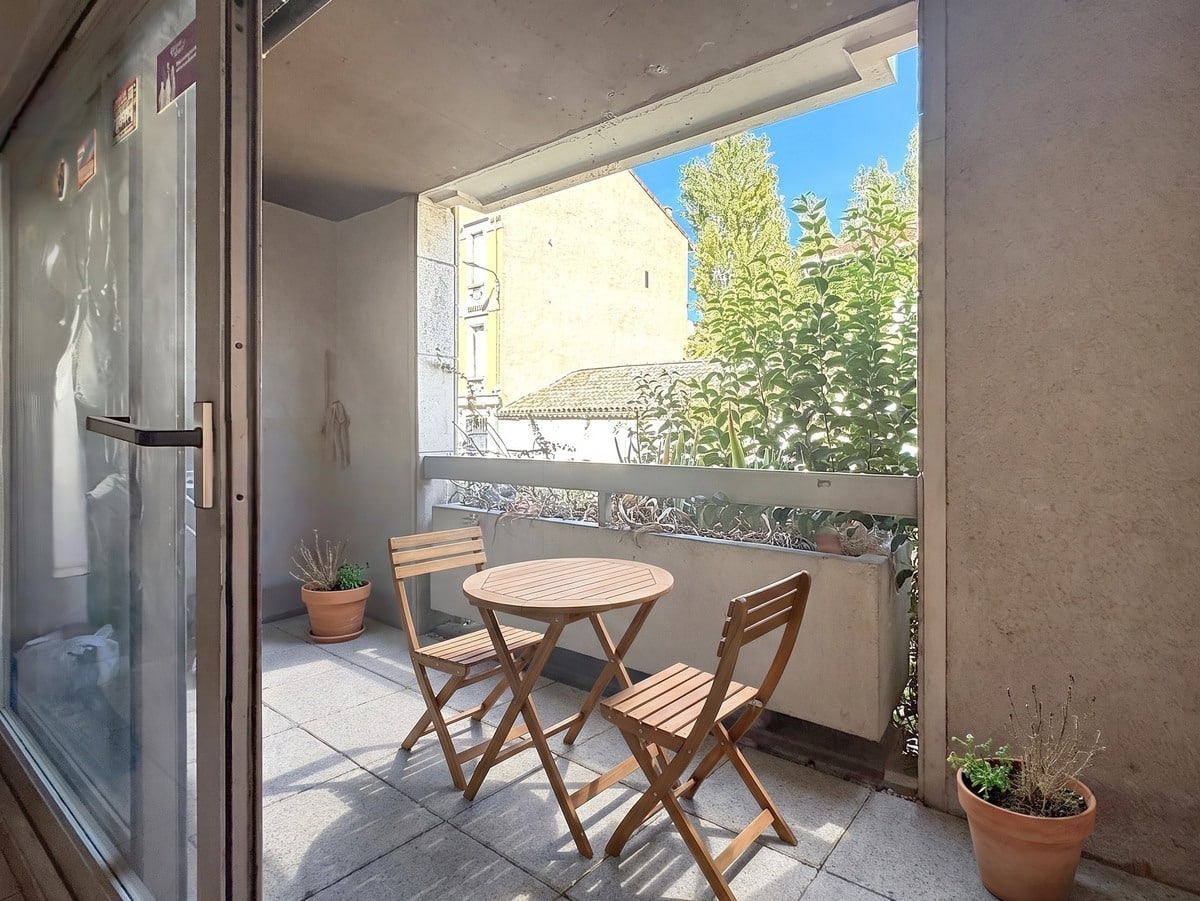 Terrasse carrelée avec une table ronde et deux chaises pliantes en bois clair, bordée d'un garde-corps en béton avec végétation abondante visible en fond sur ciel bleu. Deux pots en terre cuite contenant des plantes aromatiques encadrent l'espace extérieur, accessible depuis le séjour par une porte vitrée coulissante.