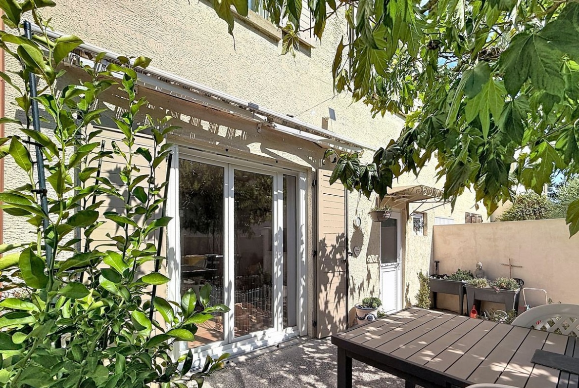 Terrasse couverte d'une pergola blanche à lames orientables avec table de jardin en bois foncé et chaises, adossée à une façade en enduit beige. Végétation dense avec arbuste à feuilles vertes en premier plan et petite porte d'accès secondaire visible sur la droite.