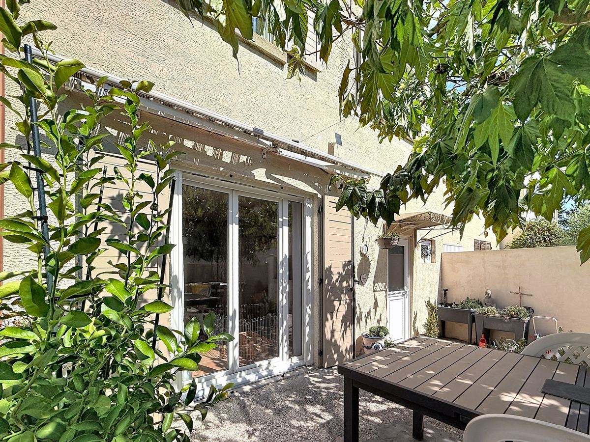 Terrasse couverte d'une pergola blanche à lames orientables avec table de jardin en bois foncé et chaises, adossée à une façade en enduit beige. Végétation dense avec arbuste à feuilles vertes en premier plan et petite porte d'accès secondaire visible sur la droite.
