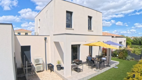 Terrasse dallée en pierre claire avec un salon de jardin composé d'une table et de chaises grises, protégée par deux parasols jaune et bleu. La maison à toiture plate avec enduit beige donne sur un jardin paysagé avec pelouse, et une unité extérieure de climatisation est visible sur le côté.