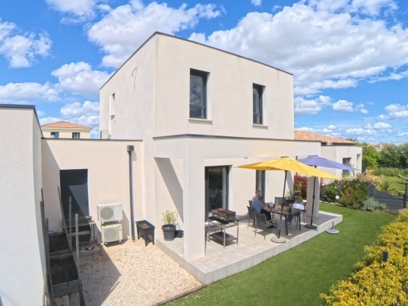 Terrasse dallée en pierre claire avec un salon de jardin composé d'une table et de chaises grises, protégée par deux parasols jaune et bleu. La maison à toiture plate avec enduit beige donne sur un jardin paysagé avec pelouse, et une unité extérieure de climatisation est visible sur le côté.