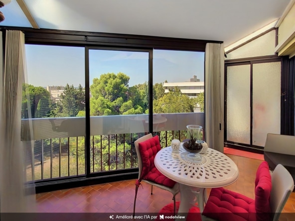 Véranda fermée par de larges baies vitrées à cadre noir offrant une vue dégagée sur une canopée d'arbres et des toits de bâtiments en arrière-plan. Un salon de jardin composé d'une table ronde blanche ajourée et de chaises à coussins rouges est installé sur un carrelage en terre cuite.