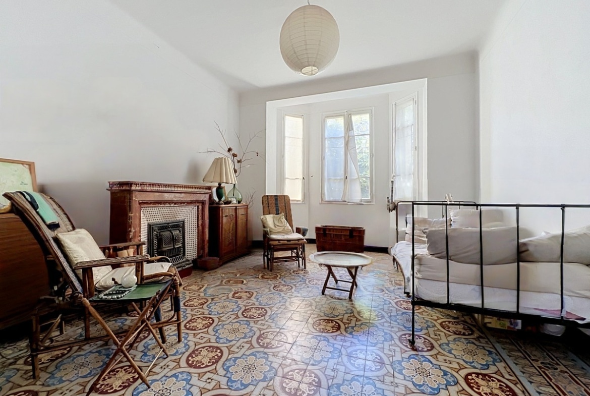 Chambre avec sol en carreaux de ciment anciens à motifs floraux polychromes, équipée d'une cheminée décorative en bois sculpté avec insert à grille métallique noire et d'un lit en fer forgé recouvert de linge blanc. La pièce dispose d'une fenêtre à trois vantaux avec petits-bois encadrée de moulures, d'une suspension boule en papier blanc au plafond et de plusieurs fauteuils en rotin.