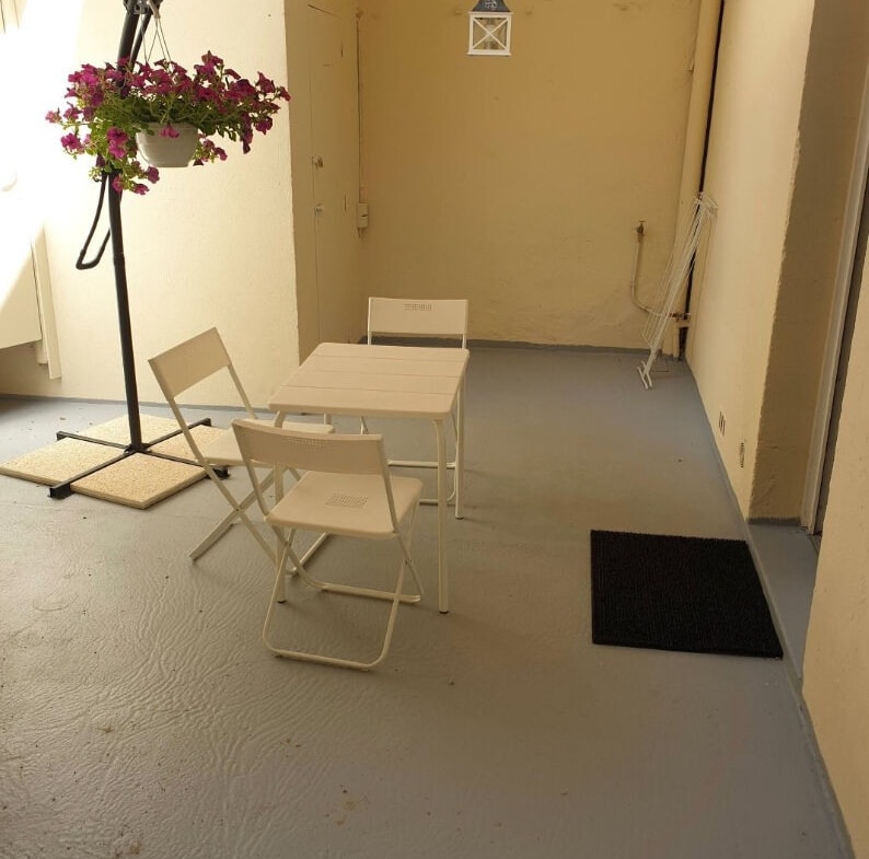 Cour intérieure privative aux murs enduits jaune pâle, sol en résine grise, avec table pliante blanche, deux chaises et parasol déporté blanc agrémenté de fleurs roses suspendues.