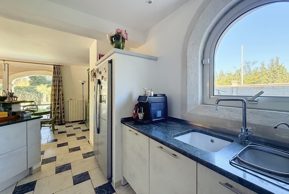 Cuisine ouverte avec façades de meubles blanches, plan de travail et crédence en granit vert foncé, évier double bac en inox sous fenêtre cintrée en pierre de taille donnant sur le jardin. Réfrigérateur américain inox et machine à café posée sur le plan de travail, carrelage damier blanc et noir au sol, ouvrant visuellement sur la salle à manger adjacente.