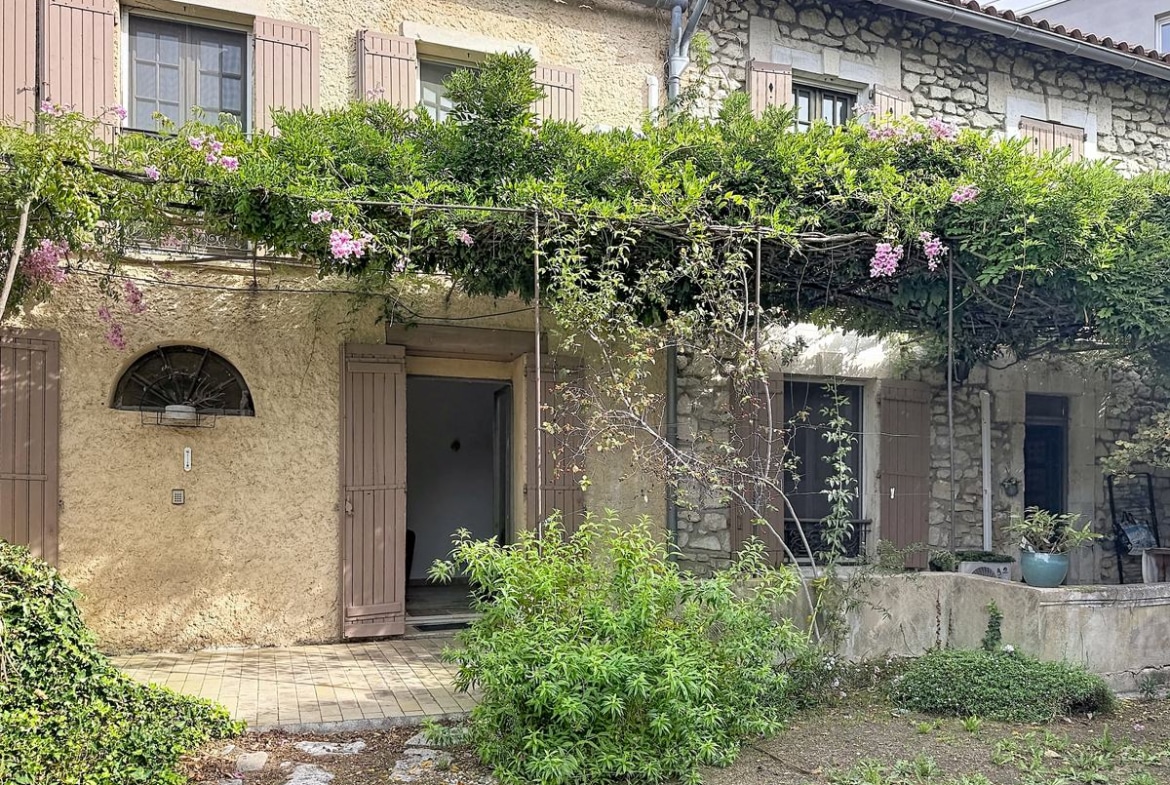 Façade principale en moellons de pierre et enduit ocre sur deux niveaux, avec volets en bois, pergola couverte de glycine et bougainvillier en fleurs roses.