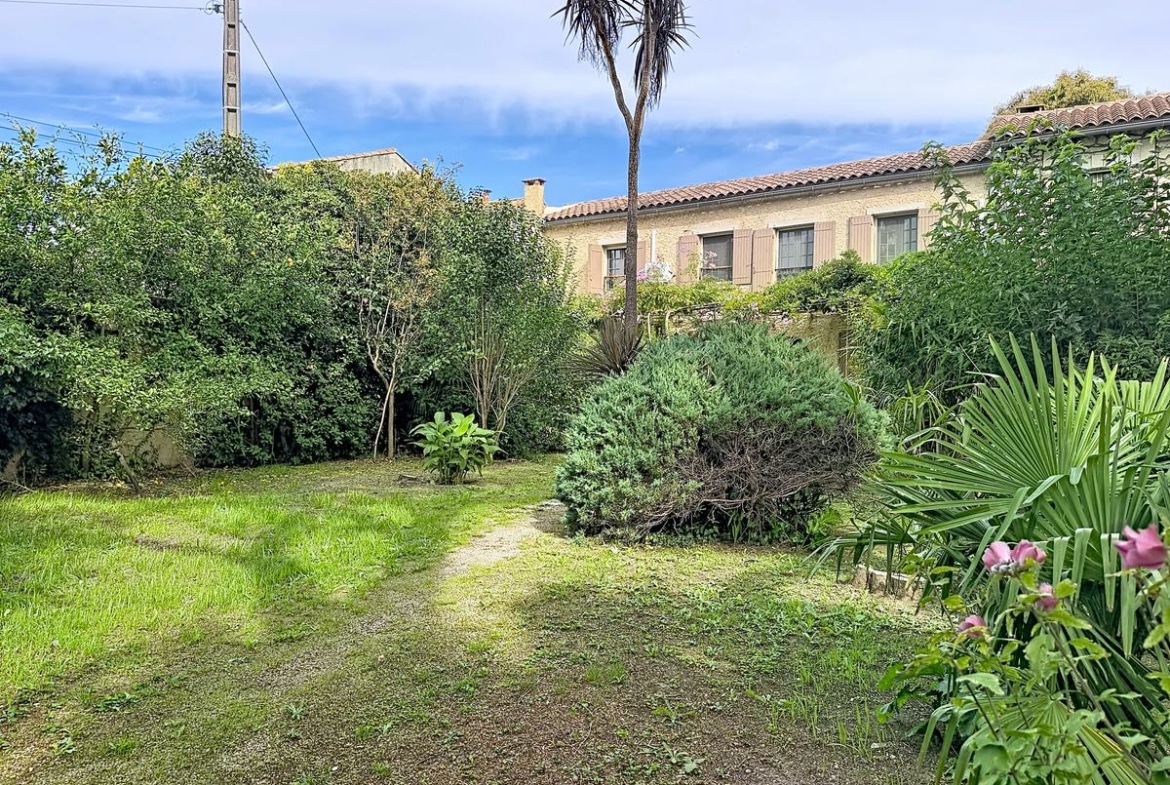 Jardin clos planté d'un grand palmier, d'arbres feuillus et d'arbustes méditerranéens variés, avec pelouse et vue sur la façade arrière du mas en pierre.