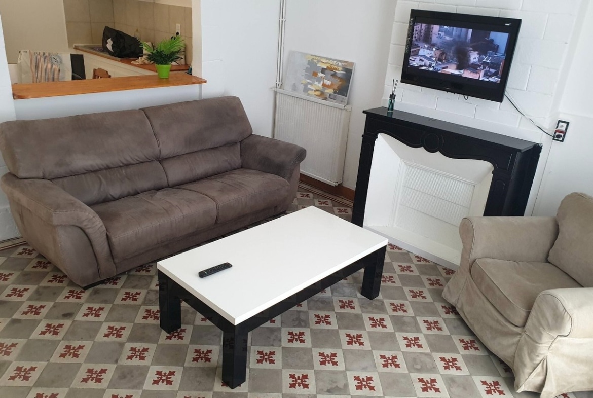 Pièce de vie avec carrelage ciment à motifs bordeaux et gris, canapé en velours taupe, fauteuil beige, table basse blanche et noire, cheminée décorative peinte en noir avec télévision murale.