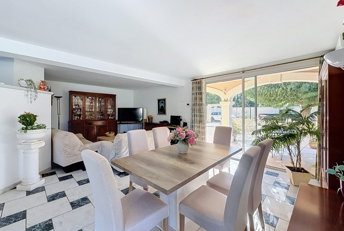 Pièce à vivre ouverte composée d'une salle à manger avec table en bois clair et six chaises tapissées beige, et d'un salon avec canapés en tissu crème, sur carrelage damier blanc et noir. Grande baie vitrée coulissante ouvrant sur la terrasse couverte à arcades, avec vue sur le jardin et la piscine.