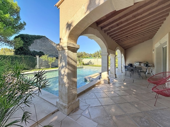 Terrasse couverte à portique de cinq arches en pierre de taille avec plafond en lambris de bois à chevrons apparents, dallage en pierres calcaires irrégulières et mobilier de jardin en fer forgé noir avec chaises rouges. Vue directe sur la piscine rectangulaire bordée de végétation méditerranéenne et sur les remparts d'Avignon visibles en arrière-plan.
